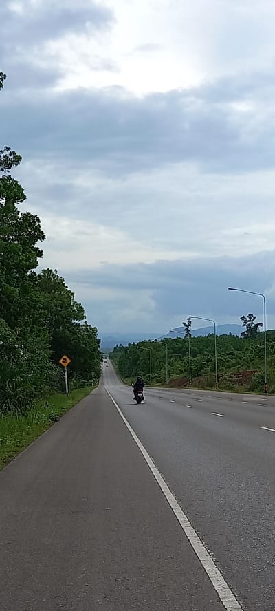 The image depicts a long, straight asphalt road in Khian Sa, Thailand, with a white line marking the edge. A motorcycle is traveling along the road. The environment includes trees and vegetation alongside the road. Streetlights are positioned at regular intervals. The sky is overcast. A road sign is visible on the roadside.