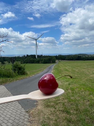 In Schwarzenborn, Germany, a single, ripe cherry is presented on a light-colored wooden spoon. The spoon is held in the foreground, with the cherry prominently displayed. The background features a winding asphalt road leading into a green, grassy field under a bright blue sky filled with fluffy white clouds. A large wind turbine stands in the middle ground, with a forest and distant hills forming the horizon. A triangular road sign is visible on the left side of the road. The overall scene depicts a rural landscape on a clear day, with the cherry serving as a focal point.