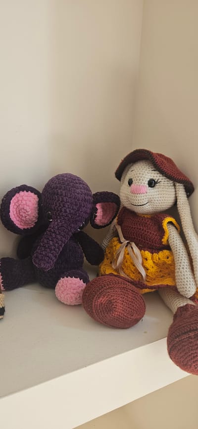 In this close-up, vertically oriented image, two crocheted stuffed animals, a bunny and an elephant, are arranged in a candid manner on a white shelf or ledge. The bunny, made of cream-colored yarn, wears a maroon bonnet and a yellow and maroon vest tied with a ribbon. Its face features black button eyes, pink rosy cheeks, and a stitched smile. The elephant, crafted from deep purple yarn, has large pink inner ears and pink foot accents. Its trunk is long and curved. The toys appear to be placed on a shelf in an indoor setting. The lighting suggests it is daytime. No text is visible in the image. The overall impression is one of whimsical display or storage of handmade toys. The location context is Hévíz, Hungary.