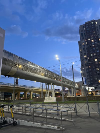 An elevated, enclosed pedestrian walkway with a curved glass exterior arches over a multi-level road system. The walkway is illuminated from within, revealing the silhouettes of people inside. A tall, modern apartment building with numerous lit windows stands to the right of the walkway. Streetlights are visible, and the sky is a deep blue with scattered clouds, indicating it is dusk. In the foreground, a metal fence separates a sidewalk from the street, and two yellow electric scooters are parked on the sidewalk. The scene is urban, with visible infrastructure like overpasses and traffic lights. The image is taken from a street-level perspective, looking upwards towards the walkway and buildings.