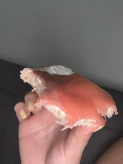 This scene, located in Albi, France, captures a human hand holding a partially consumed pastry in an indoor, dimly lit environment. The hand, featuring light yellow fingernail polish on the visible digits, grips a confection with a white interior or base and a smooth, reddish-pink glaze. The pastry exhibits irregular, torn edges consistent with recent consumption. The immediate background behind the hand is a dark, soft-textured surface, while the upper background is a plain, lighter gray wall.