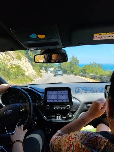 The image depicts the interior of a car driving on a road in the vicinity of Mattinata, Italy. The view is from the perspective of a passenger. In the foreground, a person's hands are visible on the steering wheel and the center console. The dashboard features a touchscreen display showing navigation and music information, with the song "Going Back to My Roots" by Richie Havens playing. The car is driving on a winding road bordered by rocky hills and lush green vegetation, with the Mediterranean Sea visible in the distance under a clear blue sky. Another car and a truck are seen driving ahead on the road. The interior of the car includes an airbag warning sticker on the sun visor and two heart-shaped stickers on the overhead console.