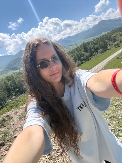 A woman is taking a selfie outdoors in a mountainous, rural area. She has long, curly brown hair and is wearing sunglasses and a light blue t-shirt with a black logo that reads "TIDE OCEAN ENDURANCE". A bright red bracelet is visible on her right wrist. The background features rolling green hills, a dirt road, a body of water, and a range of mountains under a partly cloudy blue sky. The location is identified as Bichiktu-Boom, Russia. The scene appears to be a sunny day.