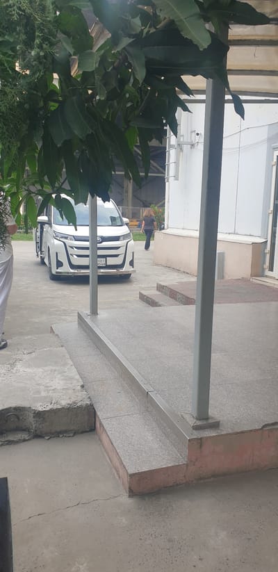 In Rooppur, Bangladesh, a white Toyota Noah minivan is parked in front of a building. A woman with long brown hair, wearing dark clothing, walks away from the vehicle towards the building. The minivan's passenger door is open. The scene appears to be outdoors, possibly near an industrial or commercial complex, with concrete paving and a ramp leading to a raised area. Trees with green leaves overhang parts of the image.
