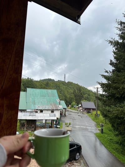 An over-the-shoulder shot captures a hand holding a green mug of coffee, with a village scene in Laništa, Montenegro, visible through a window or opening. The mug is in the foreground, obscuring part of the view. The background depicts a rural setting with a wet asphalt road, several buildings with green and grey corrugated metal roofs, a stack of firewood, a black car parked on the side of the road, and lush green trees covering rolling hills. A cell tower is visible in the distance on top of a hill. The sky is overcast and appears to be raining lightly, with several raindrops visible in the air. The scene conveys a sense of calm, possibly during a break or while observing the weather from indoors.