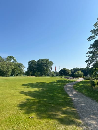 The image depicts a wide, well-maintained green lawn, likely a golf course, with a winding gravel path in the foreground. Tall, lush green trees line the background, with a clear blue sky above. Several flags, one red and two green, are visible in the mid-ground, suggesting a sports or recreational facility. In the distance, there appears to be an area with tables and umbrellas, possibly an outdoor seating area for a clubhouse. The overall scene is serene and idyllic, bathed in bright sunlight. The environment is natural and open, with no visible people or animals. This scene is consistent with the landscape of the Wassenaar Golf Club in Wassenaar, Netherlands, known for its expansive green grounds and tree-lined fairways.
