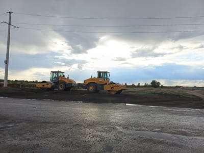 The image depicts a construction site in the city of Shakhtinsk, Kazakhstan, under a cloudy sky. Two yellow road rollers are positioned on dark soil, with a wet, grey, asphalt-like surface in the foreground. In the background, a power line pole stands to the left, and a rural landscape with vegetation and dirt roads stretches to the horizon. The overall scene suggests ongoing road construction or maintenance work.