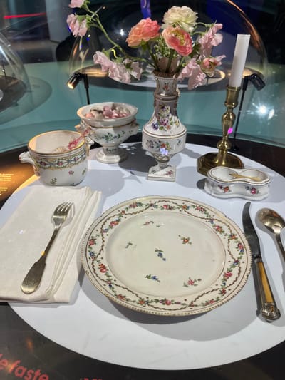This image presents a meticulously arranged antique table setting, displayed within an exhibition at the Cité Internationale de la Gastronomie et du Vin in Dijon, France. The collection of porcelain and silverware rests on a white, semi-reflective circular surface, encased behind a clear, curved barrier.
In the foreground, a white porcelain plate, adorned with a delicate floral garland border and scattered small floral motifs in its center, is positioned centrally. To its left, a silver-toned fork with a broad handle rests upon a neatly folded white napkin. To the plate's right are an ornate knife with a light, possibly bone or ivory, handle and a silver-toned spoon.
Behind the plate, from left to right, are several additional items. A white porcelain teacup, decorated with floral patterns and a patterned rim, sits to the left. Adjacent to it is a footed white porcelain bowl with a lid, containing numerous small, pale pink and white candies. Central to the rear of the display is a tall, slender white porcelain vase, richly decorated with colorful floral patterns, holding an arrangement of artificial flowers, including pink sweet peas and orange ranunculus. To the right of the vase, a brass candlestick supports a white taper candle. The furthest item to the right is a small, two-compartment white porcelain dish, likely a salt cellar, containing a miniature golden spoon in each compartment.
The background is dimly lit with hints of blue, and reflections are visible on the clear barrier. Small, focused spotlights illuminate the individual display items. Partially visible text, including "e faste," is present on a dark surface in the lower left portion of the frame, indicating an exhibit label.