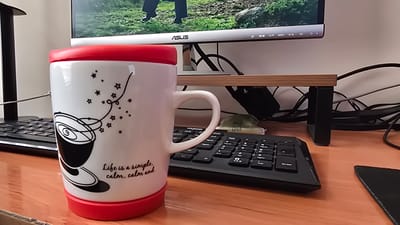 A close-up, eye-level, indoor shot captures a white ceramic mug with a red lid and base, decorated with black line art of a coffee cup and stars, and the text "Life is a simple, calm, calm and." The mug sits on a wooden desk in front of a black keyboard. Behind the mug and keyboard, a monitor displays an image of a person walking on a grassy terrain, with the ASUS logo visible on the monitor's bezel. A wooden monitor stand is positioned on the desk, with a tangle of black cables behind it. The scene suggests a workspace environment. The location of Nukus, Uzbekistan is not discernible from the image.