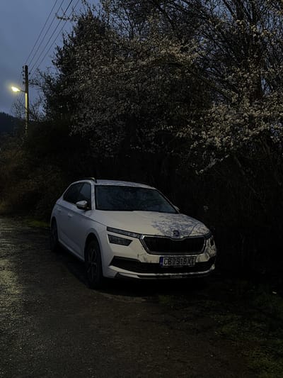 A white Skoda SUV with the Bulgarian license plate "CB 7515 XT" is parked on a dark, narrow road. The right side of the vehicle is close to the edge of the road. To the left of the car is a tree in full bloom. Overhead power lines and a street lamp illuminate the area in the distance. The location is likely near Tompsan, Bulgaria, given the license plate.