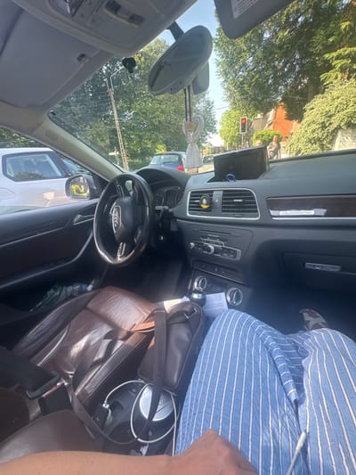 The image shows the interior of an Audi Q3, likely taken from the driver's seat. The vehicle is stopped at a red traffic light, as seen through the windshield. In front, there is a white delivery van with reflections of trees visible on its windows. The Audi's dashboard is in view, showing the infotainment screen and climate control panel. A Lacoste leather bag sits on the passenger seat, obscuring part of the blue and white striped shirt worn by the passenger. A dangling tassel-style ornament hangs from the rearview mirror. The outside environment is characterized by green trees and residential buildings, typical of a suburban neighborhood. The location is likely Marcq-en-Barœul, France, based on contextual information provided.