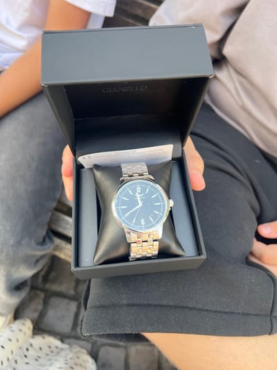 A person is holding a black watch box open, revealing a silver analog watch with a black face. The watch has a metal band and the brand name "GIANELLO" is visible on the face. The watch box has the same brand name printed vertically on the inside of its left wall. The setting appears to be outdoors, with what looks like cobblestones beneath. The person is wearing dark pants. The lighting suggests it is daytime, with sunlight creating shadows. The focus is on the watch and the box.