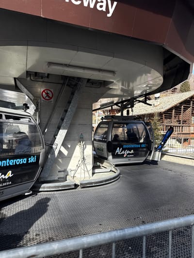 The image, correctly oriented, depicts a gondola lift station on a bright, sunny day in Alagna Valsesia, Italy. Two modern gondola cabins are prominent: one labeled "14" and another "15." Both cabins feature the branding "monterosa ski Alagna Valsesia," clearly indicating their purpose within the Monterosa ski area. Faint reflections of individuals can be seen inside the glass-enclosed cabins.
The station itself features a sturdy, multi-level structure with concrete walls, metallic support beams, and railings. The ground is covered with a dark, textured, possibly rubberized matting, likely for traction. On a brown-colored section of the station building, part of a white text reading "way" is visible, suggesting a larger sign like "Skyway" or "Gondolaway." A red circular sign with a crossed-out human figure, typically signifying a warning or prohibition, is affixed to a concrete wall.
The bright sunlight casts strong shadows across the station, suggesting it is mid-morning or early afternoon. In the background, beyond the station, there are views of buildings with stone and wooden elements, suggesting a village or other resort infrastructure, and possibly distant mountains or hills. A pair of blue skis, branded "GRACE," are seen propped against a railing, further confirming the ski resort setting. The scene captures a functional moment at a ski lift, with gondolas either waiting for passengers or moving through the station.