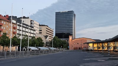 The image depicts the Jönköping Central Station area, also known as Resecentrum, in Jönköping, Sweden. In the background, a tall, modern glass building, identified by the sign "QUALITY HOTEL MATCH," dominates the skyline. To its left, a row of buildings with varied architectural styles, including a red-brick building and a white building with dark horizontal accents, are visible. In the foreground, a paved area, likely a parking lot or bus terminal, stretches out. To the right, a low-rise building with a glass facade, labeled "RESECEMTRUM," is illuminated from within, showcasing seating areas and digital displays. Several bus shelters with curved, translucent roofs are lined up along a wooden fence. Green trees are planted in a row between the buildings and the shelters. The sky is overcast with soft, diffused light, suggesting it is either early morning or late evening. There are no people or vehicles clearly visible in the scene.