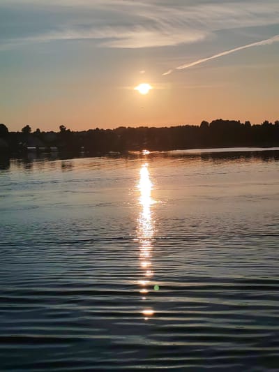 The image depicts a sunset over a body of water, likely a river or lake, with a silhouetted treeline and buildings on the horizon. The sun is visible in the upper center of the frame, casting a bright reflection on the rippling water. A contrail stretches across the sky. The foreground is dominated by the dark, textured surface of the water, with the sunset's light creating shimmering highlights. The overall mood is serene and peaceful, characteristic of a sunset scene in a natural environment. The location is identified as the city of Bol'shoi Dvor, Russia. There are no people, actions, or interactions visible in the scene.