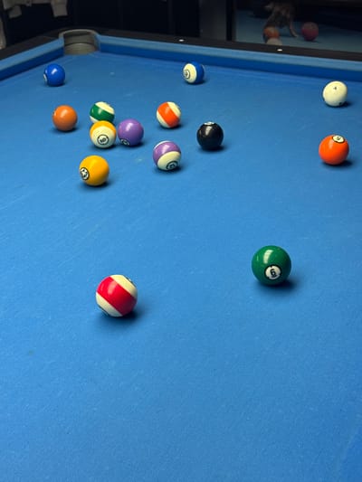 The image depicts a billiard table with multiple pool balls scattered across its blue surface. The balls are numbered and colored. The table's rail is visible along the left edge, and part of the room in which the table is located can be seen in the background. There is nothing present in the image that could be used to determine its location. The image cannot be determined to be taken in the city of Bellville, South Africa.