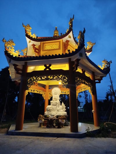A low-angle, medium shot captures a white Buddha statue centered within a traditional Vietnamese temple pavilion. The pavilion, illuminated by warm, internal lighting, has a layered roof adorned with golden dragon figures along the eaves. The structure is supported by reddish-brown pillars. Offerings, including plants and fruit, are arranged in front of the statue on a small table. The sky is a deep blue, suggesting twilight. The surrounding area appears to be a park or garden, with sparse vegetation visible in the darkness. The location is Phú An, Vietnam.