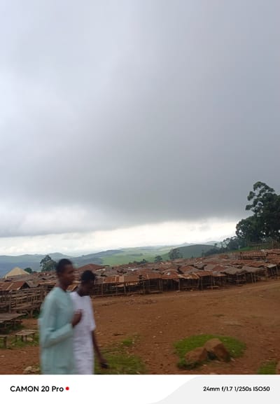 The image shows two individuals walking in a dusty area in front of a settlement with numerous structures featuring corrugated tin roofs. The settlement appears to be located on a hillside, with rolling green hills and trees visible in the background under a cloudy sky. The overall scene suggests a rural or semi-urban environment in the city of Nguroje, Nigeria.