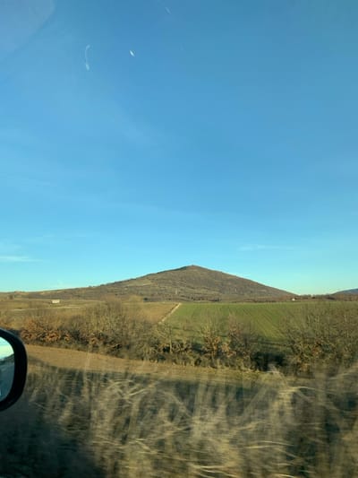 The image captures a rural landscape under a clear blue sky, likely taken from inside a moving vehicle. The primary scene is divided by a prominent horizon line. The vast upper portion of the image is dominated by a bright, cloudless blue sky, showing only a few faint, wispy marks, possibly reflections or very thin contrails.

Below the horizon, the landscape features a sloping hill or mountain range covered in what appears to be dormant or sparse vegetation and bare deciduous trees, suggesting late autumn, winter, or early spring. To the right of this hill, there is a distinct, vibrant green agricultural field, contrasting with the muted brown and grey tones of the surrounding dry land and leafless trees. A dirt track or path can be seen cutting through the midground.

The far right side of the image, extending into the foreground, displays significant light reflections and streaky patterns on a surface, consistent with a car window. A portion of a car's side mirror is visible in the bottom right corner, confirming the perspective from inside a vehicle, possibly in motion.

The overall impression is of a bright, clear day in a mountainous agricultural area. No people are visible. No explicit text is discernible within the image. The setting aligns with the geographical characteristics of Grevena, Greece, known for its mountainous terrain and rural landscapes.