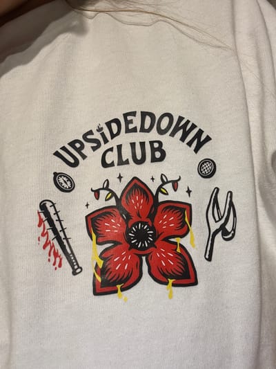 The image is a close-up of a white crew neck sweatshirt with a graphic design. The design features the words "UPSIDEDOWN CLUB" in a curved, black, sans-serif font. Below the text is a red, flower-like creature with black petals and a dark center. Yellow liquid drips from the creature. To the left of the creature is a baseball bat with blood splatters. To the right of the creature is a pair of black tongs or pliers. Above the creature, to the left, is a compass-like symbol, and to the right is a round object with a textured surface. The sweatshirt appears to be worn by a person with blonde hair visible at the top of the frame. The background is out of focus and appears to be a plain surface. There is no indication of the location being in Getafe, Spain.