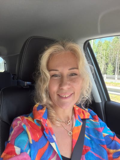 The media file is a close-up selfie of a woman taken inside a car. The woman has blonde, wavy hair and is smiling at the camera. She is wearing a colorful, abstract-patterned shirt with shades of blue, orange, pink, and yellow. A gold necklace with two interlinked rings is visible around her neck. The background shows the interior of a car, with black leather seats and a portion of the car's interior ceiling. Through the car window, a glimpse of trees and a blue sky can be seen, suggesting an outdoor setting and daytime. There is also some text visible on the window, which appears to be "SUZUKI" and "E1" which are likely car manufacturer and model related. The overall mood is cheerful and casual. The location context provided is Rokhma, Russia.