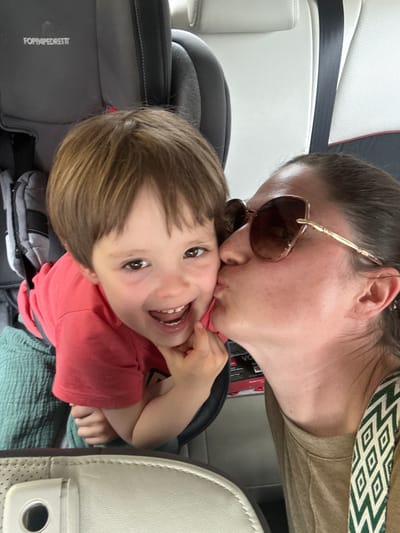 A woman wearing sunglasses is kissing a young child on the cheek. The child is smiling and has a hand near their face. They are both in what appears to be a car seat inside a vehicle. The location of Meljine, Montenegro, cannot be confirmed from the image.