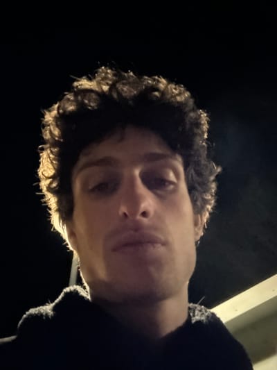 A single male individual is depicted in a close-up, low-angle shot, directly facing the camera. The scene is set at night in Riorges, France. The subject has dark, curly hair and is wearing a dark, textured hooded garment. A prominent light source is positioned behind and to the left of the individual, creating a distinct rim light around his hair and left shoulder. The front of his face is largely shadowed due to the backlighting. The background is predominantly dark and indistinct, with a faint, light-colored structural element visible in the lower right portion of the frame.