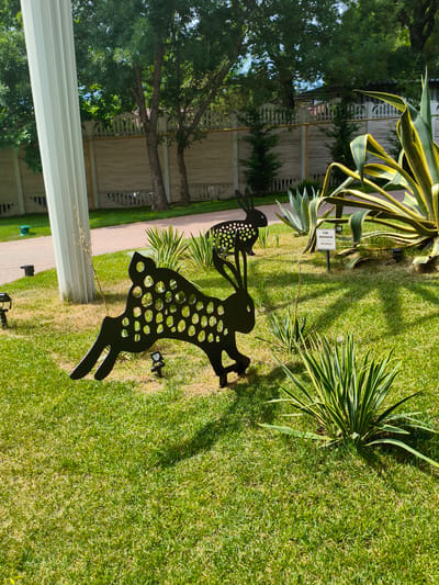 This image captures a sunny outdoor scene in Glebovskoye, Russia, likely a park or garden area. The foreground is dominated by a well-manicured green lawn. Two black, silhouetted metal sculptures of rabbits are prominently placed on the grass. The larger rabbit in the foreground is in motion, with its legs extended, and features a pattern of circular cutouts across its body. A smaller rabbit sculpture, also with circular cutouts, is positioned behind and slightly above the larger one.
In the background, a light-colored, ornate fence runs horizontally across the frame, separating the garden from a pathway and trees beyond. A large, white fluted column stands to the left of the frame, partially obscuring the fence. Various plants are visible, including a tall agave plant with variegated leaves in the middle ground to the right, and smaller, spiky plants scattered throughout the lawn. Two small, dark garden lights are visible near the rabbit sculptures. A white sign with text in Russian is placed in front of the agave plant. The sky is visible through the leafy canopy of trees in the upper portion of the image. The overall impression is of a decorative garden setting on a clear day.