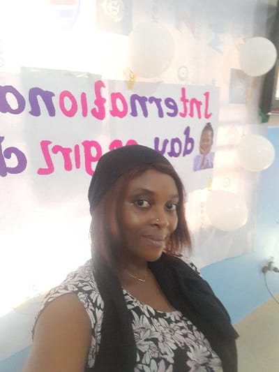 The image depicts an indoor scene in Ajah, Nigeria, centered on a female individual. She is positioned in the foreground, slightly right of center, looking directly at the camera with a mild smile. The woman has medium-brown skin, reddish-brown hair partially covered by a dark headwrap, and a small silver-colored nose ring on her right nostril. She is wearing a dark-colored top with a white floral pattern and a dark shawl draped over her shoulders. A thin gold-colored necklace with a small V-shaped pendant is visible around her neck.
The background features a white banner or sign with mirrored text, indicating "International Day of the Girl Child." A small image of a young girl is visible on the banner, to the right of the mirrored "Day" text. Several white, spherical balloons are affixed to the wall or banner in the background. The wall behind the banner appears light-colored, transitioning to a light blue section on the far right, where a dark electrical outlet is visible. The overall lighting is bright and even.