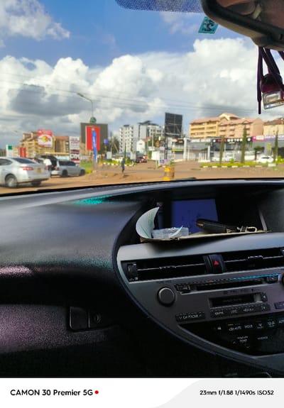 The image is taken from the driver's seat of a car, looking out towards a busy street scene in Kiambu, Kenya.  The street is lined with buildings and shops, with a few pedestrians and vehicles visible. The sky is cloudy and bright, with some clear patches of blue sky. There are several buildings visible in the background, including a large building with a sign that says "City Motors." The car's interior is black and grey, with a black dashboard. There are several objects visible on the dashboard, including a cell phone, a key chain, and a small box.  The car is facing forward, and the image captures a scene of a typical city intersection, with the driver's perspective.  The coordinates -1.203, 36.834 suggest that the scene is near the intersection of Limuru Road and Kenyatta Road in Kiambu.  The car's dashboard is in focus, while the exterior scene is slightly blurred.  The camera lens is a 23mm lens, and the photo was taken with a 1/1490 second shutter speed and an ISO setting of 52.