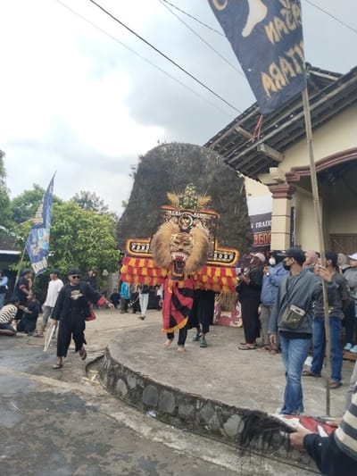 Captured in Pendem, Indonesia, the image shows a traditional cultural performance taking place outdoors, with a crowd gathered around. The central focus is a large, elaborate headdress resembling a lion's head with a mane of feathers and colorful embellishments. Performers hold the headdress aloft, moving along a stone platform. Other performers and spectators stand nearby, some wearing casual clothing and face masks. Banners and flags are present, adding to the festive atmosphere. The surrounding environment includes a house with a traditional roof, trees, and a paved area.