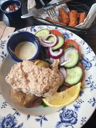 A close-up, eye-level shot displays a meal on a decorative white plate with blue floral patterns. The meal consists of a baked potato topped with a generous portion of tuna mayonnaise, a side salad with tomatoes, cucumbers, and red onion, a wedge of lemon, and a small blue bowl of salad dressing. In the background, another blue bowl holds ketchup, and a black basket contains several pieces of fried fish sticks, with a fork and knife resting on white paper above them. The setting appears to be a restaurant or pub in Epsom and Ewell, United Kingdom, suggested by the style of the food and presentation. The lighting is natural and ambient, highlighting the textures and colors of the food.