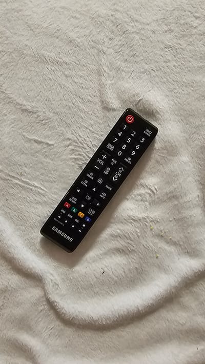 A black Samsung television remote control lies on a fluffy white surface. The remote is angled diagonally with the top left corner towards the top right of the frame. Visible text on the remote includes numbers one through nine and zero, a power button icon, and labels such as "SOURCE," "PRE-CH," "MUTE," "VOL," "CH," "GUIDE," "INFO," "RETURN," "EXIT," "REC," "SETTINGS," "AD/SUBT.," and colorful buttons labeled A, B, C, and D. The brand name "SAMSUNG" is printed vertically along the bottom right edge of the remote. The white surface appears to be a soft fabric, possibly a blanket or towel, with subtle shadows and folds, suggesting it's likely indoors. There are a few very small yellow specks and a couple of dark specks on the white surface, but no other objects or people are visible. The lighting appears to be artificial, casting soft shadows.