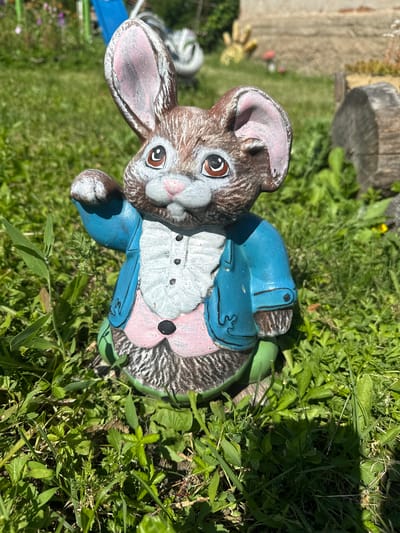 A ceramic garden statue of a rabbit, painted with brown fur, pink inner ears, a white ruffled shirt, a blue jacket, and a pink vest, is positioned in the center of the frame. The rabbit is seated, facing the viewer, with its right arm raised in a waving gesture. Its eyes are open and looking forward, and its mouth is slightly open. The statue is placed on a patch of green grass with scattered weeds and some sunlight casting shadows. In the background, out of focus, are glimpses of a blurry blue structure, a piece of weathered wood, and some foliage. The setting appears to be an outdoor garden. There is no discernible connection to Belo pole, Bulgaria.