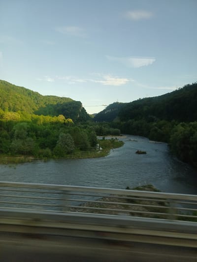 The image, rotated 90 degrees counter-clockwise, captures a serene natural landscape featuring a river flowing through a valley flanked by forested hills under a clear sky. The river occupies the central and right portions of the scene, its waters appearing a deep blue-gray, with small sections of rocky or vegetated banks and islets visible within its course. Lush, dense green trees cover the slopes of the hills, suggesting a rich forest environment. The sky above is a bright, light blue with a few scattered white, wispy clouds.

In the distance, on the higher parts of the left hill, faint structures resembling power lines or utility poles can be seen, indicating a subtle human presence within the expansive natural setting. The immediate foreground on the right side of the image is dominated by vertical metal bars, likely part of a railing or fence, and a blurred, darker panel, suggesting the photograph was taken from a window, possibly of a moving vehicle like a train, or from a bridge or elevated structure.

There are no people visible in the scene. The clear, bright lighting indicates it is daytime, likely morning or afternoon, with good weather conditions. The overall impression is one of peaceful natural beauty. The location context provided is Galitsyno, Russia.