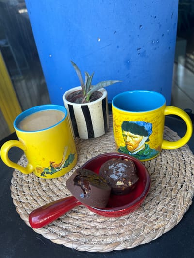 A close-up, overhead shot features a collection of items arranged on a woven straw placemat. The dominant subjects are two bright yellow mugs with blue interiors, each adorned with a painted portrait. One mug is filled with a light-colored beverage. To the left of the mugs, a small red dish holds two pieces of chocolate confections and a spoon with a red handle. Behind the red dish, a small potted plant with striped black and white ceramic pot is visible. The items are placed on a woven straw placemat with a criss-cross pattern, which rests on a dark surface, likely a table. To the right of the placemat is a bright blue surface, possibly a table or countertop, with some visible water droplets. The scene appears to be outdoors, with natural light suggesting daytime. The overall mood is casual and domestic. No visible text is present in the image.