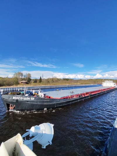 A barge named "VOLGO-DON 101" is moving through a canal. The barge is black with red accents and a gray deck. There are trees and some buildings on the canal bank. The sky is blue with white clouds. The image appears to have been taken from another vessel. The location is Анхимово, Russia.