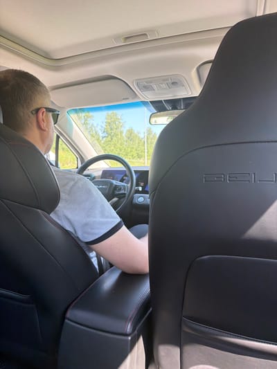 In the city of Zapadny, Russia, a man is pictured from behind as he drives a BYD car. He is wearing sunglasses and a grey t-shirt with black trim. The interior of the car is visible, including the dashboard, steering wheel, and a large digital display screen. The car has a sunroof, which is open, allowing sunlight to filter into the cabin. The seats are black leather with red stitching. The background shows a bright, sunny day with green trees and blue sky, suggesting the car is on a road outside of a dense urban area.