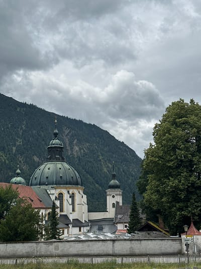 The image depicts Ettal Abbey, a Benedictine monastery located in the Bavarian Alps near Ettal, Germany. The main structure features a large, green, domed roof with ornate detailing and a spire topped with a golden cross. To the left of the main dome, another smaller green dome is visible. The building is white with yellow accents and has several arched windows. A tall, white bell tower with a green, pointed roof rises to the right of the main dome.
In the background, a steep, tree-covered mountain dominates the skyline under a cloudy, grey sky. A large, lush green tree stands in the foreground on the right side of the image, partially obscuring the view of the abbey and the mountain. A grey stone wall runs across the middle ground, with a wooden fence and some dry grass in front of it. Several smaller buildings, possibly part of the monastery complex, are visible behind the wall, some with red roofs and others with glass structures. A street sign with directional arrows is attached to a small, red-roofed structure on the far right. The overall atmosphere is one of a somber, overcast day in a mountainous, historic setting.