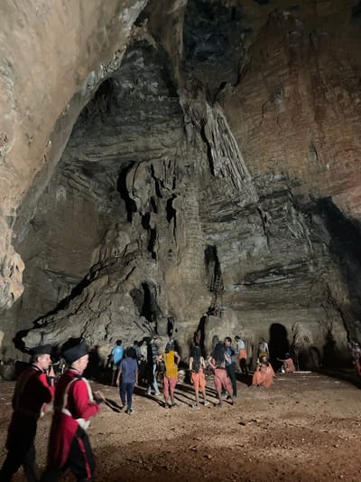 The image depicts the interior of a large cave, likely a tourist attraction, in Gollagutta, India. The cave features a high, arched ceiling with prominent stalactites and natural rock formations. The floor is covered with reddish-brown dirt and gravel.
In the foreground, two individuals are dressed in red and black historical costumes, possibly representing guards or performers. They are facing away from the camera, moving towards the center of the cave.
Further into the cave, a group of people are gathered. Some are dressed in what appear to be traditional Indian clothing, including brightly colored patterned tops and draped garments in orange and yellow hues. Other individuals are wearing more casual attire, such as t-shirts and trousers. One person is seated on the ground in an orange robe. A ladder is visible on a rock face in the background, suggesting there might be an elevated area within the cave or access to other parts.
The lighting is dim, indicating the natural light is limited, with artificial lighting illuminating the scene, casting shadows on the cave walls and floor. The overall impression is that of a guided tour or a staged event within the natural cave environment.