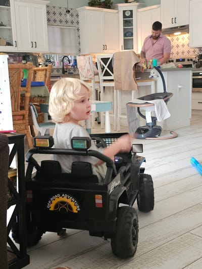 The image depicts an indoor scene, likely a kitchen and living area, possibly in Islamorada, United States of America. A young child with blonde curly hair is seated in a black toy Jeep. The child is wearing a grey t-shirt and looking to their right with their mouth slightly open. The toy Jeep has "OFF-ROAD ADVENTURE" written on its rear. In the background, a man wearing a pink collared shirt is standing at a kitchen counter, appearing to prepare food. The kitchen features white cabinets, a tiled backsplash, and a stovetop. A baby swing is visible near the counter. The flooring appears to be light-colored wood. The overall lighting suggests daytime.