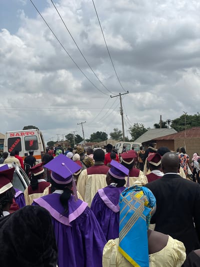 A procession of people, some wearing graduation gowns and caps, moves along a street in Kassa, Nigeria. The group includes individuals in purple and maroon academic regalia, as well as people in civilian attire. In the background, a white van with "NARTO BAWASA" written on its side is visible, along with utility poles and buildings lining the street. The sky is overcast with clouds.
