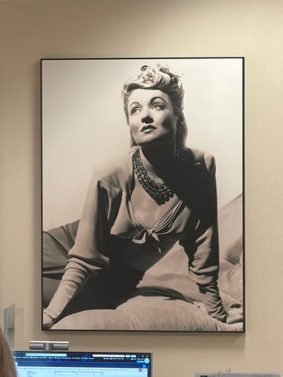 A framed black and white portrait of a woman, likely from the mid-20th century, hangs on a beige wall. The woman has her hair styled up with a flower accent, wears a necklace, and a knotted blouse with full sleeves. She is reclining on a cushioned surface. The bottom of the frame is visible above a computer monitor and other office equipment, suggesting the location is an office environment. The image appears to be a photograph, possibly an artistic print. No specific landmarks of Burbank, United States of America are visible in this image.
