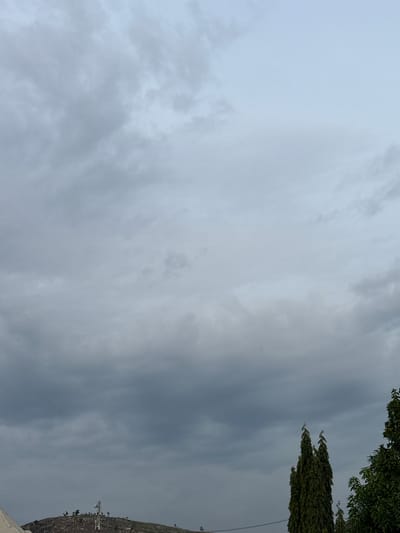 The image depicts a slightly overcast sky over a rocky, elevated landscape. In the foreground, the tops of several tall, slender trees are visible on the right, with their dark green foliage contrasting against the muted sky. A portion of what appears to be a white structure is present in the lower left corner. Dominating the mid-ground on a rocky hilltop is a telecommunications tower, a white structure with multiple platforms and antennas. Scattered trees with dark green foliage are also present on the hilltop. The sky is filled with varying shades of gray and white clouds, suggesting a diffused light or an approaching weather change. The scene appears to be a rural or semi-rural area within Angwan Sayawa, Nigeria, characterized by its natural topography and the presence of infrastructure for communication. There are no people or animals visible in the frame.