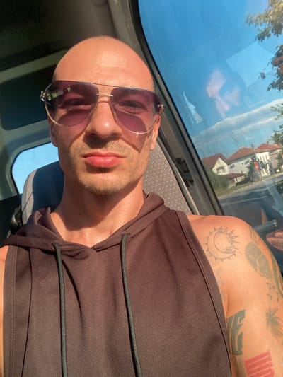 The media file shows a close-up, low-angle, tilted shot of a bald man wearing sunglasses and a dark brown tank top. He has stubble on his face and appears to have tattoos on his arm, including a crescent moon with rays and a silhouette of a bird. The man is indoors, possibly in a vehicle, with a window to his left reflecting the sky and some buildings with red roofs. Sunlight is casting a warm glow on his face and arm. The scene suggests it might be daytime, with the sun shining. The overall emotion conveyed is relaxed, perhaps due to the sunglasses and casual attire. The location is contextually identified as Petrevene, Bulgaria. There is no visible text in the image.