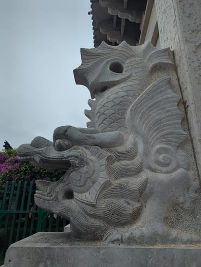 This media file features a close-up, low-angle shot of intricately carved stone ornamentation, likely part of a traditional Chinese architectural element. The carving depicts a mythical creature, resembling a dragon or a Qilin, with scales, elaborate fins, and a fierce, stylized head. The detail in the carving is evident, showcasing textured scales, flowing mane-like features, and sharp teeth. The stone itself appears weathered, suggesting it is an older structure.
The background is dominated by a hazy, overcast sky, indicating a cloudy or foggy day. Below the stone carving, a glimpse of lush green foliage with vibrant purple flowers can be seen, suggesting an outdoor setting with vegetation. A dark, slatted fence is also partially visible in the background. The perspective is from below, looking upwards at the architectural detail, which is mounted on a larger stone structure. The overall lighting is soft due to the overcast sky. There is no visible text in the media file. The scene evokes a sense of historical artistry and cultural significance.