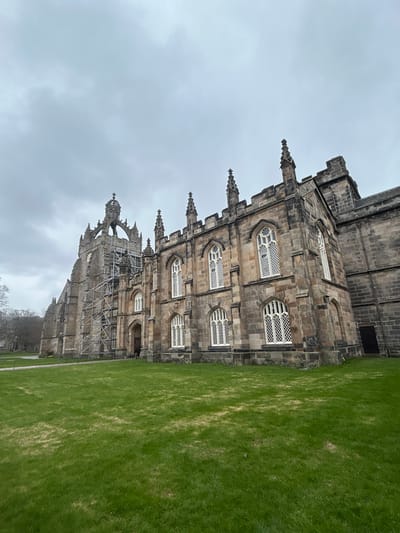 The image shows a large, ornate stone building with Gothic architectural elements, including spires, pointed arches, and decorative windows. The building is likely a historic structure, possibly a university or church. Scaffolding is erected on one section of the building, indicating ongoing maintenance or restoration work. The building is adjacent to a grassy lawn. The sky is overcast, suggesting a cloudy day. The location is Aberdeen City, United Kingdom.