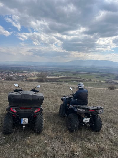 Two dark-colored All-Terrain Vehicles (ATVs) are positioned on a dry, brown-yellow grassy hillside overlooking the city of Balsha, Bulgaria, and its surrounding valley.
The ATV on the left is parked with its rear facing the camera, featuring a large black storage box on its rear rack. Its Bulgarian license plate reads "CO A BG 5033." The ATV on the right is also parked, with a single individual seated on it, facing away from the camera towards the distant landscape. The person is wearing a dark blue jacket, dark trousers, and a white and black helmet. This ATV's license plate reads "C 25687." Both ATVs have knobby tires suitable for off-road terrain.
In the mid-ground, the village of Balsha is visible with numerous houses featuring red-tiled roofs, interspersed with green fields and sparse trees. Beyond the valley, a prominent mountain range with snow-capped peaks (consistent with Vitosha Mountain) extends across the horizon. The sky is partly cloudy, with patches of blue visible amidst large grey and white clouds. The overall environment suggests a rural, elevated vantage point.