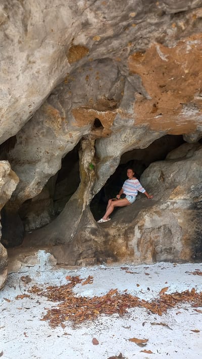 A middle-aged woman in a blue, white, and red striped shirt and denim shorts is standing inside the mouth of a cave with white sandals. She appears to be leaning into the cave with her left hand touching the rock. The cave is located in the city of Thap Put, Thailand. The background is composed of cave walls and rock formations. The ground outside the cave is white, covered with dry leaves and debris. The image appears to be taken from a slightly low angle.