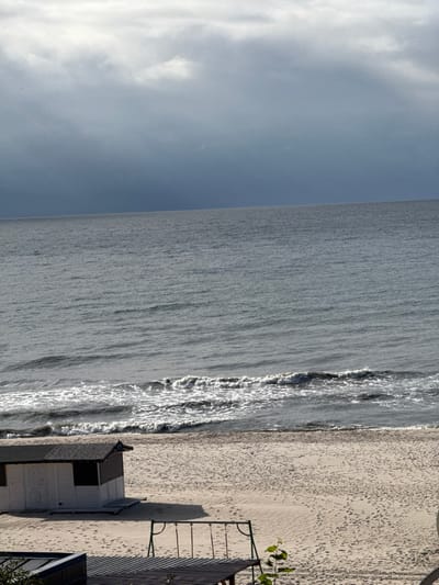 The image displays a coastal scene in Yantarny, Russia, under an overcast sky. The upper half of the frame shows dense, grey cloud cover, darkening towards the horizon line where it meets the sea. The Baltic Sea occupies the middle portion of the image, appearing grey-blue with visible ripples and several lines of white-foamed waves breaking near the shore. Below the sea is a wide expanse of light tan sand, marked with numerous footprints. On the sandy beach, positioned towards the mid-left, is a small, single-story, white building with a dark, sloped roof and a white door, casting a distinct shadow to its right. Further into the foreground, on the right side of the beach, a greenish-brown metal swing set frame with three suspended swings is visible, also casting a shadow. The bottom left foreground includes a dark, angular structure, possibly a part of a roof or deck, and a small green plant. No individuals are present in the scene.
